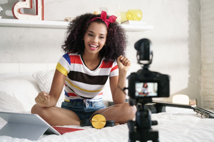 “Medium-dark-skinned influencer talks to her SLR camera on her bed, promoting a lead magnet to her audience.”